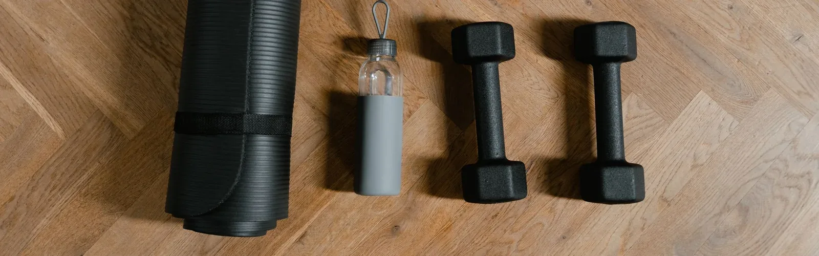 Two black hex dumbbells beside a water bottle and the end of a rolled mat on a wooden floor.